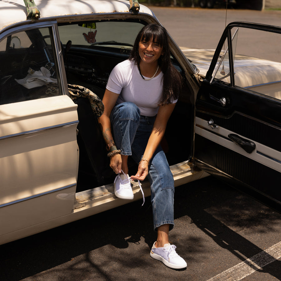Woman tying left-foot Reef Neptune sneaker facing forward, seated in a vintage car, showcasing beach-inspired comfort footwear.