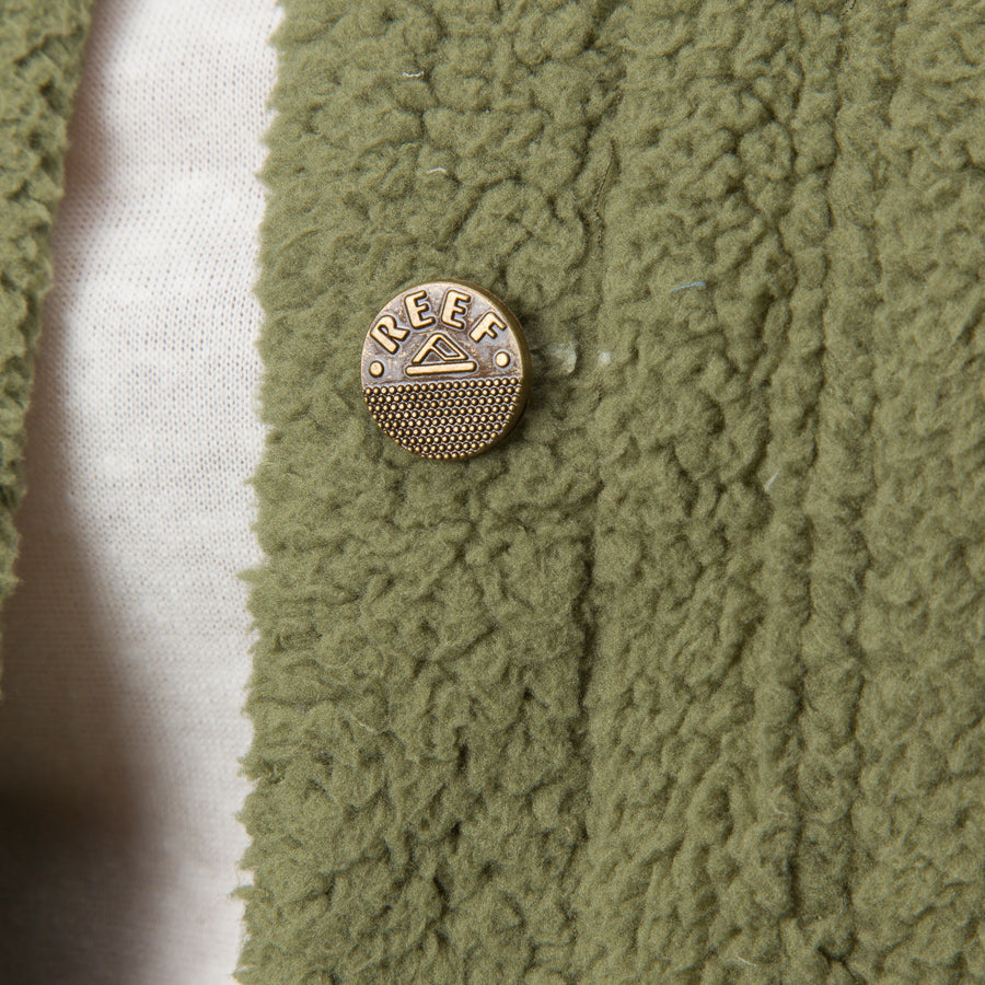 Close-up of olive green Sammie Sherpa Jacket fabric showing texture and REEF metal logo button for comfort-focused beach apparel.